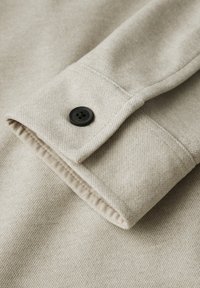 Tejido de sudadera beig con acabado texturizado, que presenta un puño doblado y un botón negro. La tela muestra un sutil patrón ligero.