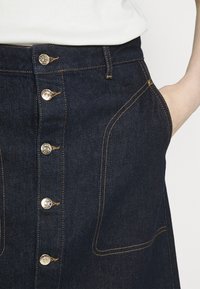 Jeanskjol i mörkblått med knappstängning fram och kontrasterande orange sömmar. Har sidofickor och en strukturerad passform.