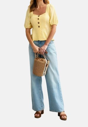 Mujer con blusa amarilla de mangas abullonadas con botones, vaqueros de pierna ancha azul claro y sandalias marrones de plataforma, sosteniendo un bolso redondo tejido.