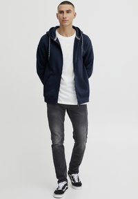 Navy zip-up hoodie met een trekkoord capuchon, twee voorkeurszakken, gedragen over een wit T-shirt, gecombineerd met donkere jeans en zwarte sneakers.