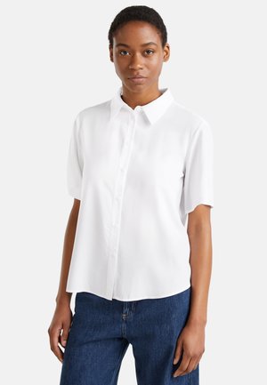 Mujer con una camisa blanca de manga corta abotonada y vaqueros azules, de pie frente a un fondo claro y sencillo.