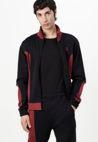 Zwarte sportjack met bordeauxrode accenten. Heeft een hoge kraag, ritssluiting en geribde manchetten. Draagt over een zwart shirt.