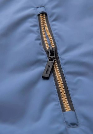 Tissu bleu clair avec une fermeture éclair noire et des dents beiges. Le tirette de la fermeture éclair est étiquetée "OMBRE." Le matériau est lisse, avec une texture minimale.