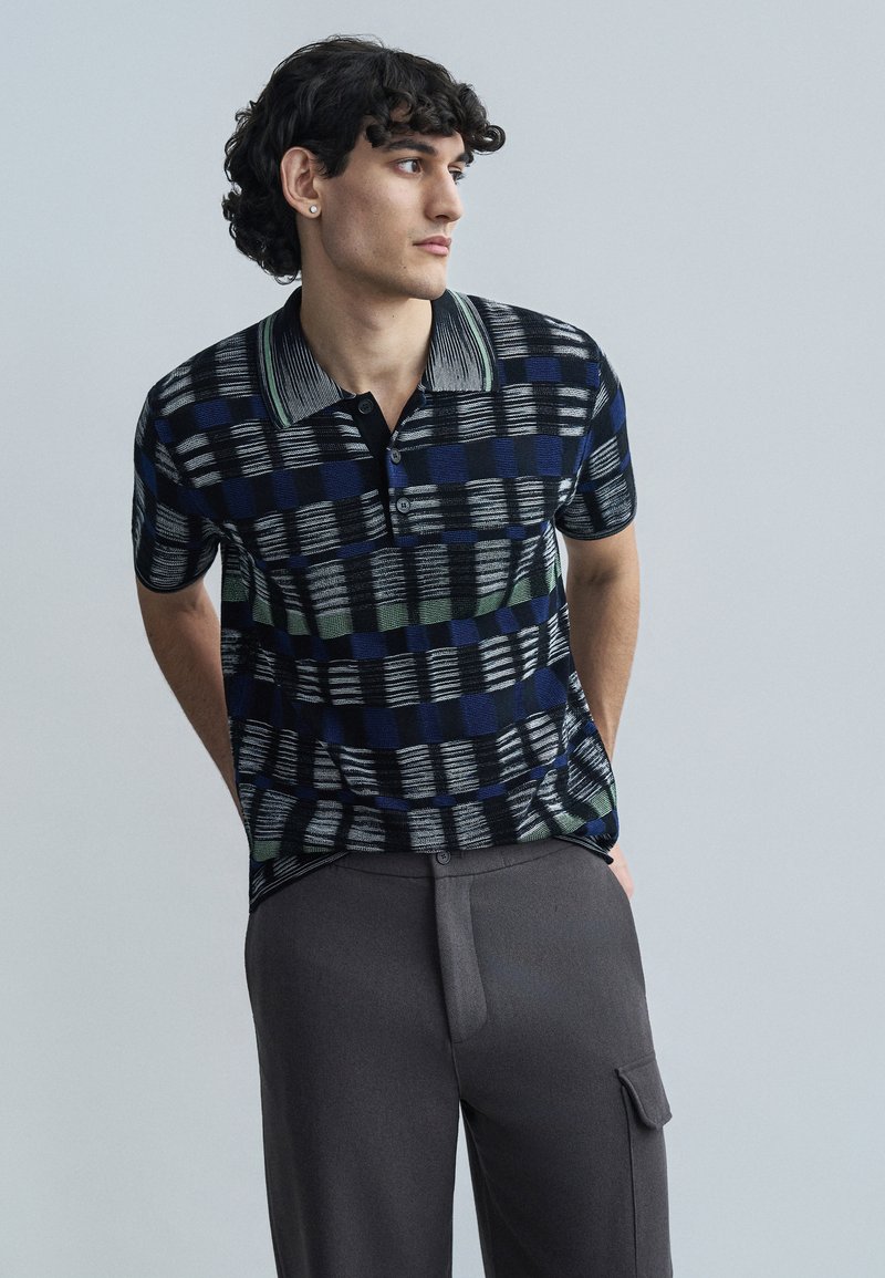 Black and blue checkered polo shirt with a striped collar and three buttons. Paired with gray trousers featuring side pockets.