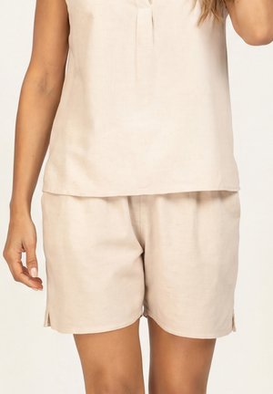 Kobieta w beżowej bluzce bez rękawów i dopasowanych, luźnych szortach na neutralnym tle, widoczna tylko w części talii i ramion.