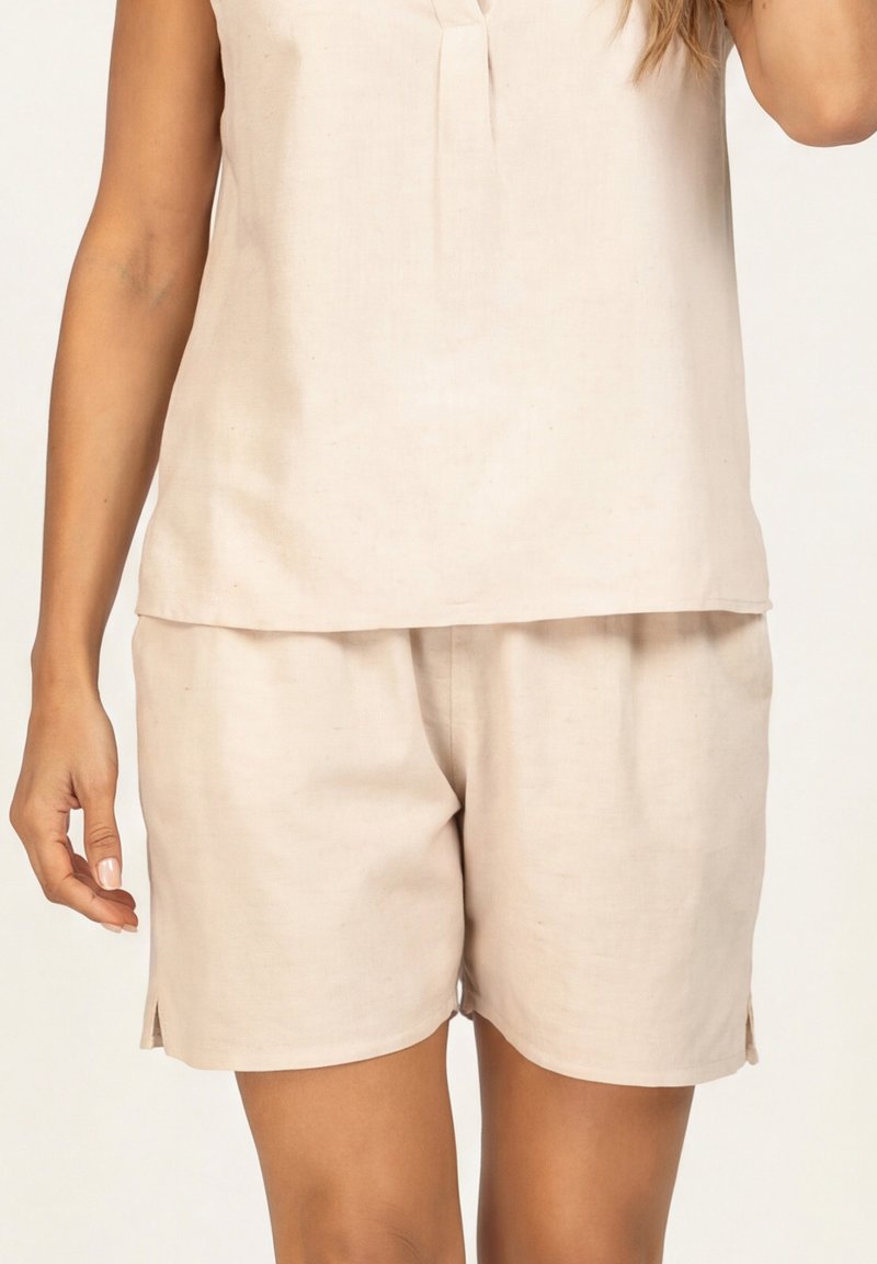Kobieta w beżowej bluzce bez rękawów i dopasowanych, luźnych szortach na neutralnym tle, widoczna tylko w części talii i ramion.