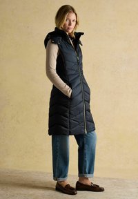 Sieviete, kas ģērbusies garā melnā quiltētā vesti ar kapuci virs gaiši brūna džempera, ar locītiem zilu džinsu piedurkņiem un brūnām mokasīnām, stāv ar rokām kabatās.