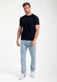 Tamsiai mėlynas megztas t-shirt su trumpomis rankovėmis ir horizontaliomis tekstūros raštais, derinamas su šviesiai mėlynomis džinsomis ir baltais sportiniais bateliais su juodais akcentais.