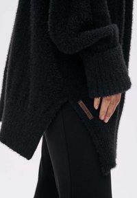 Zwarte oversized trui van fuzzy materiaal, met een zijsplit en een bruin merkembleem op de zoom. Gecombineerd met zwarte broek.