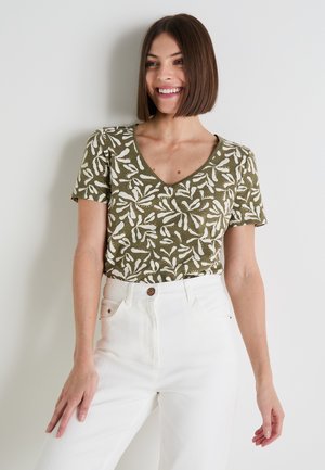 Camiseta verde oliva con patrón de hojas blancas, escote en V y mangas cortas abullonadas, combinada con pantalones de mezclilla blancos de talle alto.