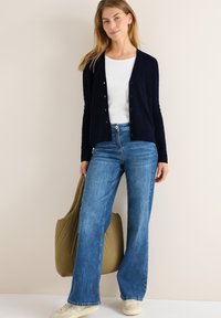 Marinblå stickad cardigan med knapplås, bärs över en vit topp. Matchas med ljusblå vida jeans och en grön axelväska. Beiga sneakers.