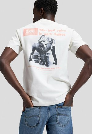 Mann in blauer Jeans steht mit dem Rücken zur Kamera und trägt ein weißes T-Shirt mit einer Vintage-Lee-Arbeitskleidungswerbung, die zwei Männer auf Fässern und Ziegeln sitzend zeigt.