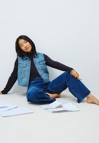 Gilet en denim sur un haut noir à manches longues, associé à un jean bleu à jambes larges. Personne assise sur un tapis clair, entourée de papiers éparpillés.