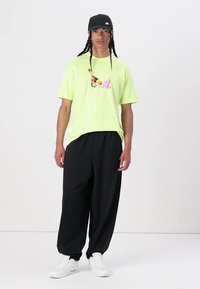 Neonově zelené tričko s grafickým potiskem uprostřed, spárované s volnými černými kalhotami a bílými teniskami. Černá kšiltovka dokončuje outfit.