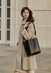 Zwarte leren tote bag met een gestructureerde vorm, voorzien van gouden hardware en een bijpassende beige jas met een hoge kraag.