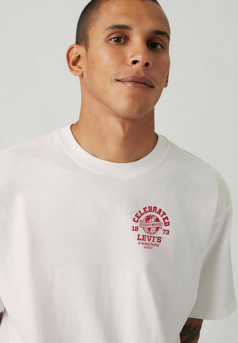 Mand med kort hår iført en hvid Levi's t-shirt med rød "Celebrated Everywhere 1873"-logo på brystet, mod en ensfarvet baggrund.