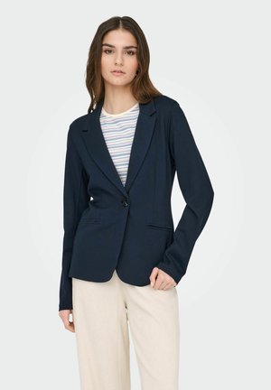 Kvinde iført en marineblå blazer med en enkelt knap over en stribet skjorte og lyse beige bukser, stående mod en ensfarvet lys baggrund.