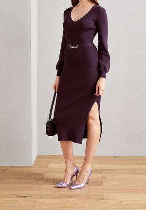 Kobieta w długiej, prążkowanej fioletowej sukience z rozcięciem, z czarnym paskiem, trzymająca małą czarną torebkę, ubrana w metaliczne fioletowe szpilki.