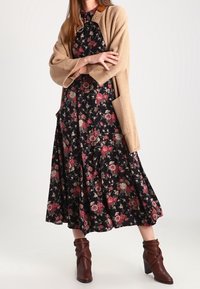 Donna in piedi con le braccia incrociate, indossa un vestito midi floreale nero, un cardigan in maglia beige e stivali alla caviglia con tacco marroni.