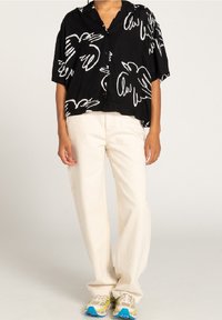 Maglietta nera a maniche corte con motivi astratti di uccelli bianchi, abbinata a pantaloni larghi color crema e sneakers multicolori.