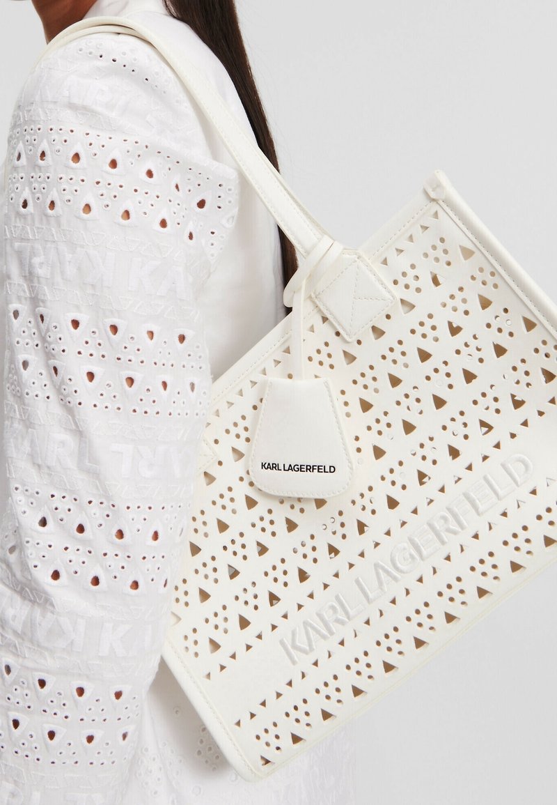 White perforated tote bag with triangular cutouts, leather material, logo tag, and structured design. Features a textured surface and long handle.
