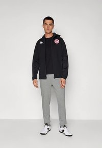 Mann, der eine schwarze Zip-Jacke mit Sportlogos, ein schwarzes T-Shirt, graue Jogginghosen und weiße Sneaker trägt, steht vor einem schlichten Hintergrund.