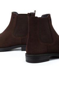Bottines Chelsea en suède marron avec des panneaux élastiques sur les côtés, une languette et des semelles en caoutchouc. Le design présente une texture lisse et un style minimaliste.