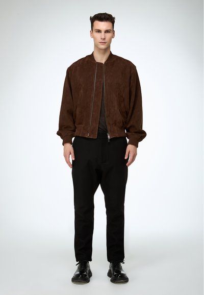 Junger Mann stehend, trägt eine braune Wildleder-Bomberjacke, schwarze Hose und schwarze glänzende Schnürschuhe vor einem schlichten hellen Hintergrund.