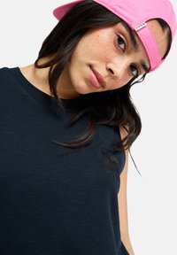 Μπλε ναυτικό αμάνικο μπλουζάκι με ριμπ υφή, που φέρει μια μαύρη κεντημένη λογότυπο στο στήθος· συνοδεύεται από ζωηρή ροζ καπέλο.