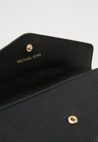 Sac en cuir noir avec rabat ouvert, montrant un bouton-pression doré et "MICHAEL KORS" embossé en or à l'intérieur.