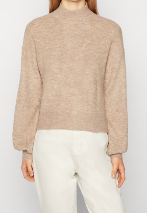 Femme portant un pull en maille côtelée beige avec de longues manches bouffantes et un pantalon taille haute blanc cassé, debout devant un fond uni.