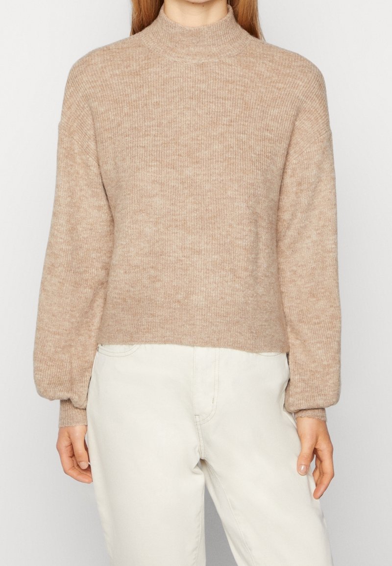 Femme portant un pull en maille côtelée beige avec de longues manches bouffantes et un pantalon taille haute blanc cassé, debout devant un fond uni.