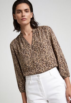 Frau trägt eine braun-schwarze Bluse mit Leopardenmuster und Dreiviertelärmeln sowie hoch taillierte weiße Hose vor einfachem Hintergrund.