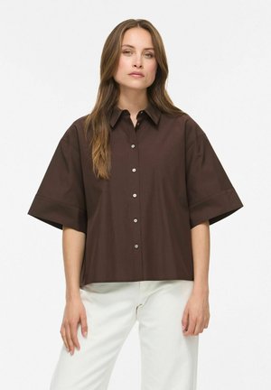 Femme aux longs cheveux bruns portant une chemise ample marron foncé à boutons et un pantalon blanc, debout devant un fond blanc uni.