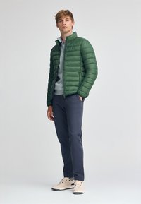 Grön vadderad jacka med horisontellt quiltat mönster, grå sweatshirt under, marinblå byxor och beige sneakers med svarta sulor.