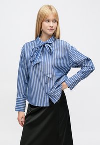 Mėlynos ir baltos spalvos dryžuota marškinėliai su sagomis, dideliu kaspinu prie apykaklės, ilgomis rankovėmis ir šiek tiek trumpintu apačia. Juodas satininis sijonas.