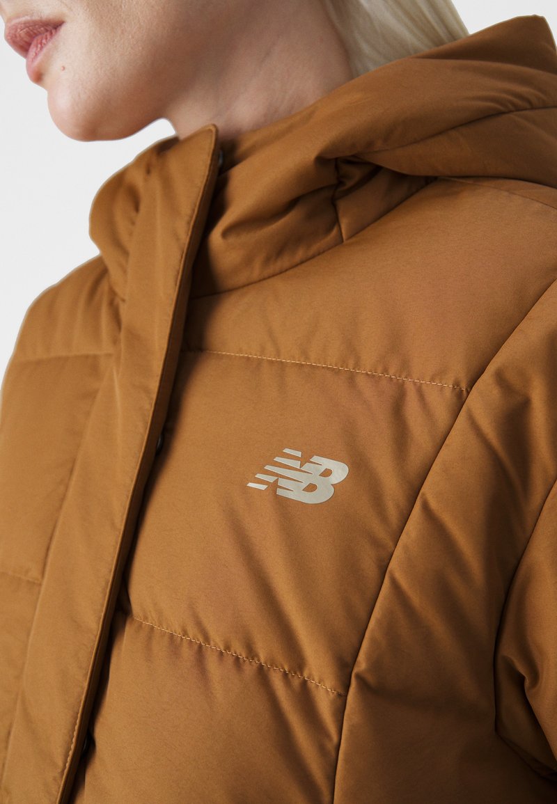 Doudoune brune avec un col haut et un logo New Balance argenté. Tissu lisse et texturé avec des coutures visibles et un détail de capuche.