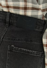 Černé džínové kalhoty s džínovou kapsou s třepícím se detailem a obdélníkovou černou koženou nášivkou s nápisem "vytvořte sebe sama." Částečně viditelná kostkovaná košile.