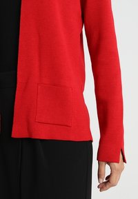 Cardigan rosso lavorato a maglia con apertura frontale, tessuto testurizzato, una singola tasca frontale e spacchi laterali. Indossato sopra top e pantaloni neri.