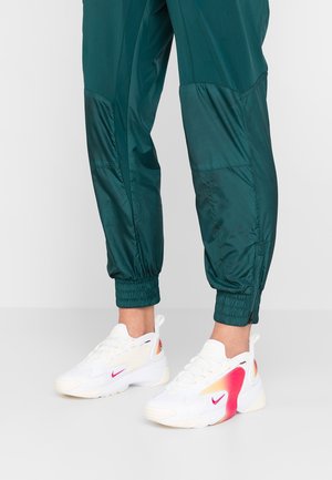 Pieds portant des baskets Nike blanches avec des accents roses et orange, associées à un pantalon de jogging bleu sarcelle avec des poignets élastiques.