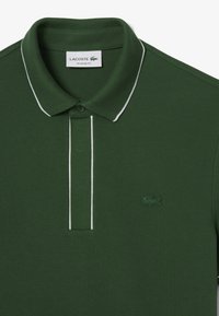 Polo di colore verde scuro, con un colletto classico bordato di bianco, tre bottoni e un logo di coccodrillo ricamato in verde sul lato sinistro del petto.