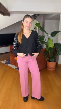 Ženska z dolgimi lasmi v čop, oblečena v črno srajco z gumbi in belimi vzorci ter rožnatimi hlačami, stoji v notranjosti blizu rastlin in pohištva.