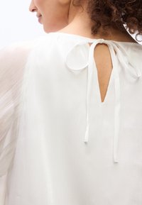 Γυναίκα με σγουρά μαλλιά που φοράει λευκό διάφανο πουκάμισο δεμένο με φιόγκο στη λαιμόκοψη στο πίσω μέρος, αποκαλύπτοντας μια μικρή οπή σαν κλειδαρότρυπα.