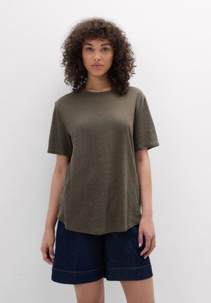 Frau mit lockigem Haar, die ein olivgrünes Kurzarmshirt und dunkelblaue Jeansshorts trägt und an einer weißen Wand steht.