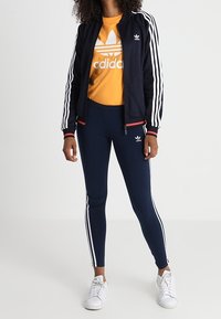 Marinhållet träningsjacka med vita ränder, orange t-shirt med logotyp och marinblå leggings med ränder. Vita sneakers fulländar outfiten.