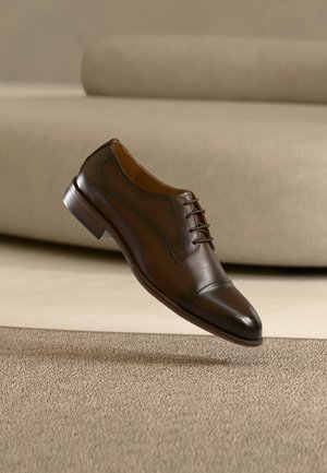 Chaussure de ville marron en cuir pour homme avec lacets flottant au-dessus d'un tapis beige et d'un canapé assorti en arrière-plan.