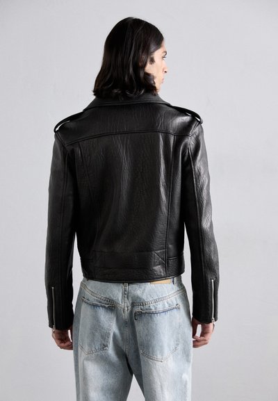 Black leather motorcycle jacket with a textured finish, shoulder epaulets, and zipped cuffs, worn over light blue denim jeans.