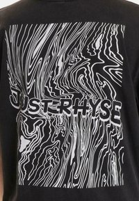Tricou negru cu un model abstract alb în spirală și text bold "JUST RHYSE". Mâneci scurte, decolteu rotund, material din bumbac.