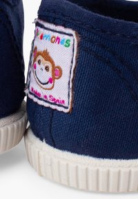 Nahaufnahme der Ferse eines dunkelblauen Stoffschuhs mit weißer Gummisohle und einem bunten Etikett mit Affengesicht mit der Aufschrift „Playshones Made in Spain.“