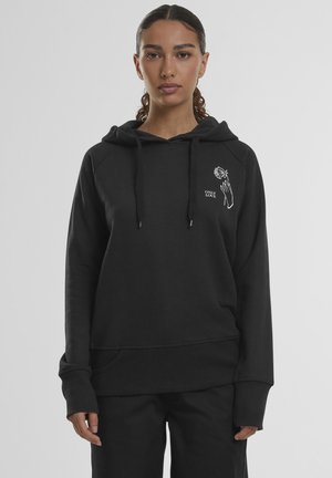 Mladá žena s vlasmi stiahnutými dozadu nosí čiernu mikinu s nápisom „ONLY LOVE“ a obrázkom ruky držiacej ružu na hrudi.